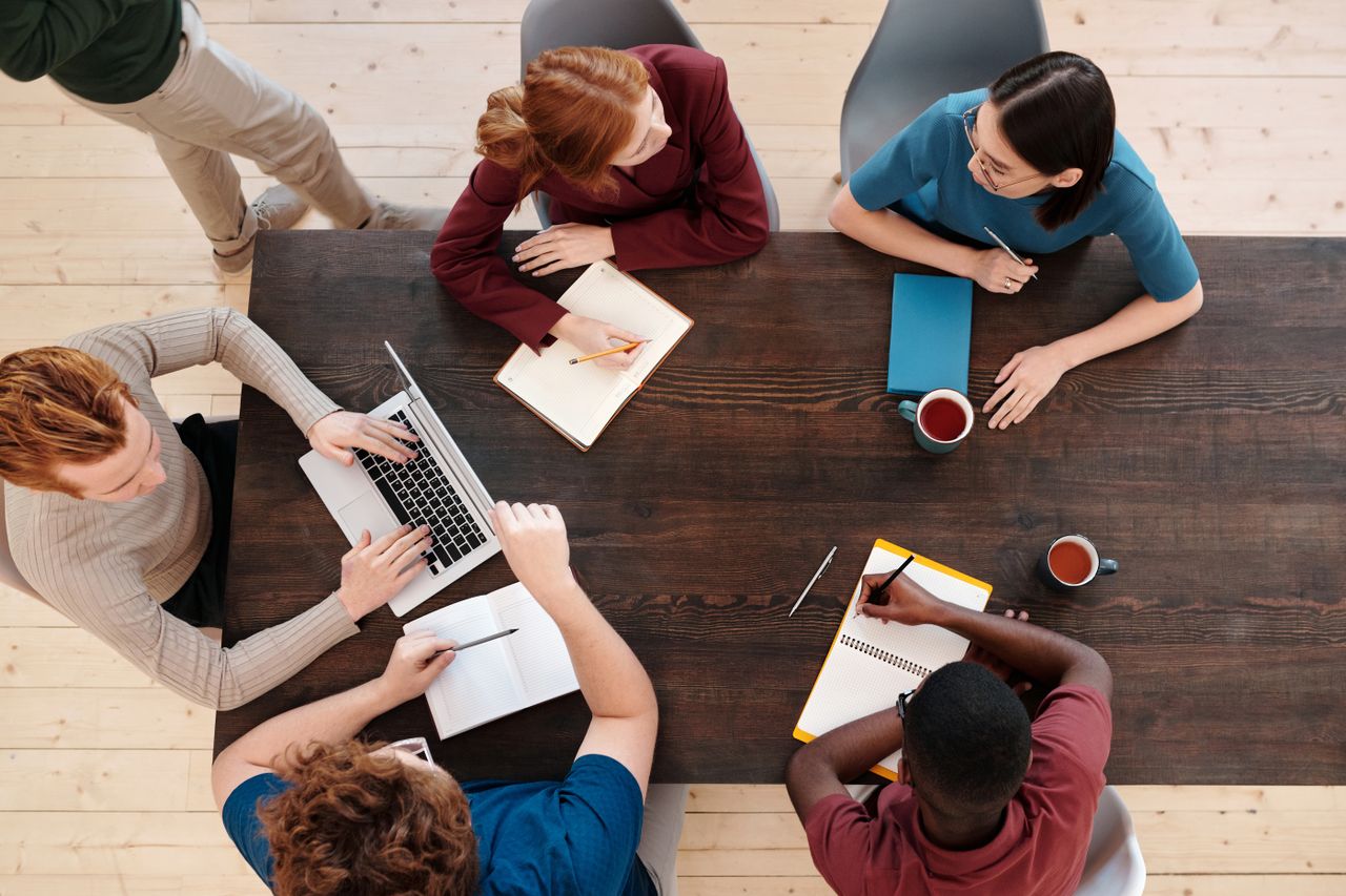 A birds-eye view of a table with people sat around with books, laptops and pens working together