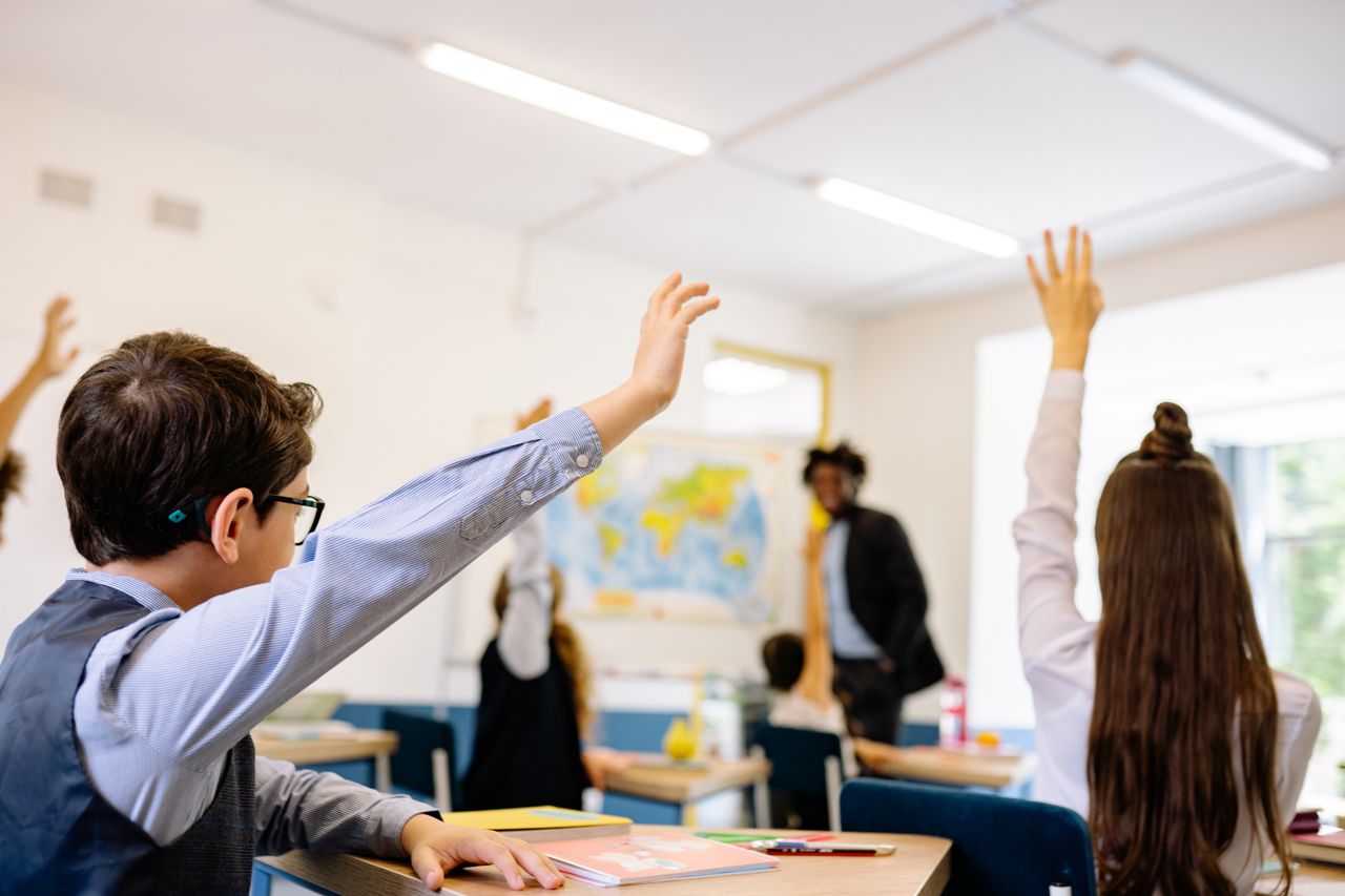 A photograph of a classroom with two students in the foreground with their hands raised with a teacher in the background